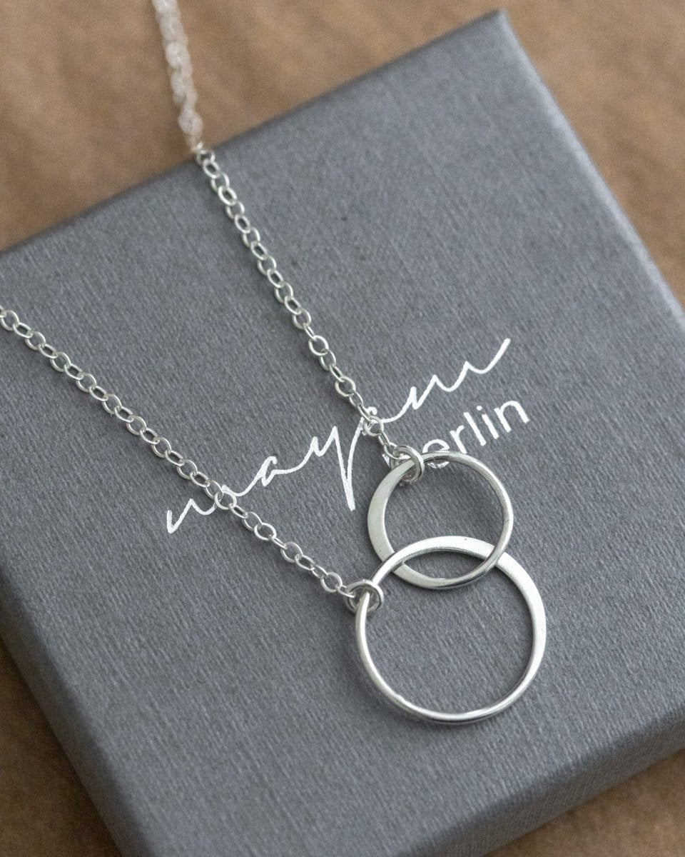 Eine zierliche 925 Silberkette mit zwei miteinander verschlungenen und unterschiedlich großen Ringen in der Mitte liegt auf einem grauen Schmucketui.