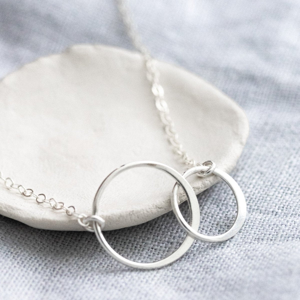 Eine zierliche 925 Silberkette mit zwei miteinander verschlungenen und unterschiedlich großen Ringen in der Mitte lehnt an einem weißen Schälchen.