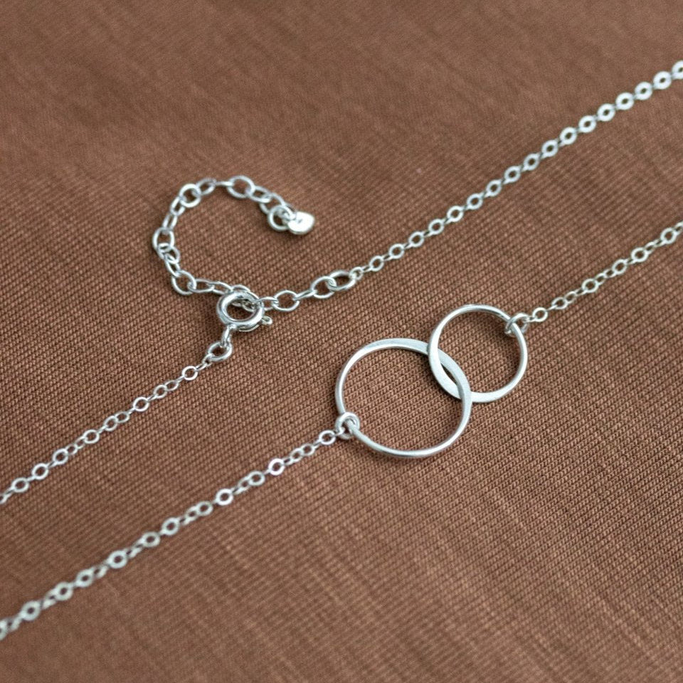 Eine zierliche 925 Silberkette mit zwei miteinander verschlungenen und unterschiedlich großen Ringen in der Mitte liegt auf braunem Stoff.