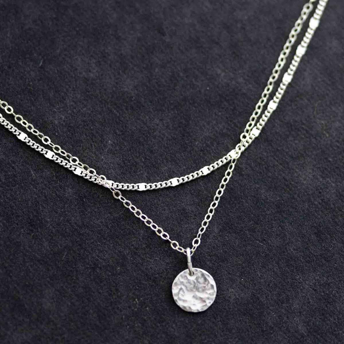 Ein Halsketten-Set bestehend aus einer kürzeren Gliederkette und einer längeren Ankerkette mit gehämmerten Plättchen-Anhänger vor dunklem Hintergrund.