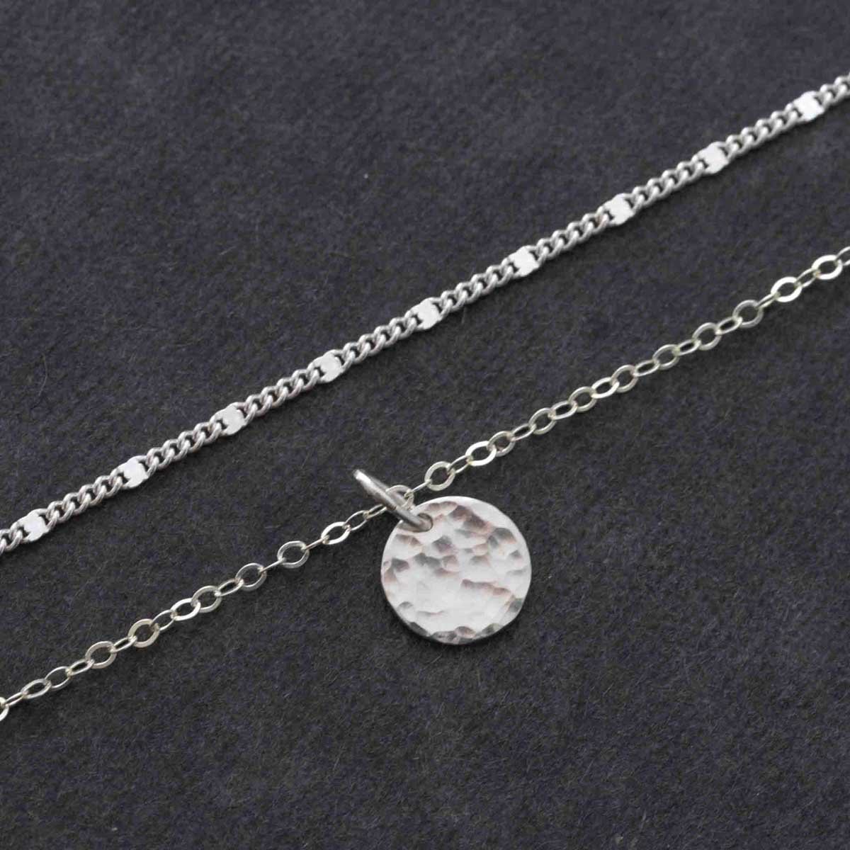 Detail-Aufnahme eines Halsketten-Sets bestehend aus einer kürzeren Gliederkette und einer längeren Ankerkette mit gehämmerten Plättchen-Anhänger vor dunklem Hintergrund