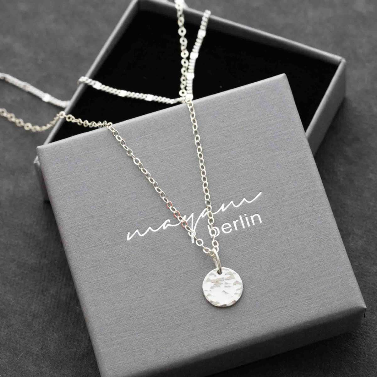 Ein Halsketten-Set bestehend aus einer kürzeren Gliederkette und einer längeren Ankerkette mit gehämmerten Plättchen-Anhänger liegen auf einem grauen Schmucketui mit den eingeprägtem Namen mayam berlin.