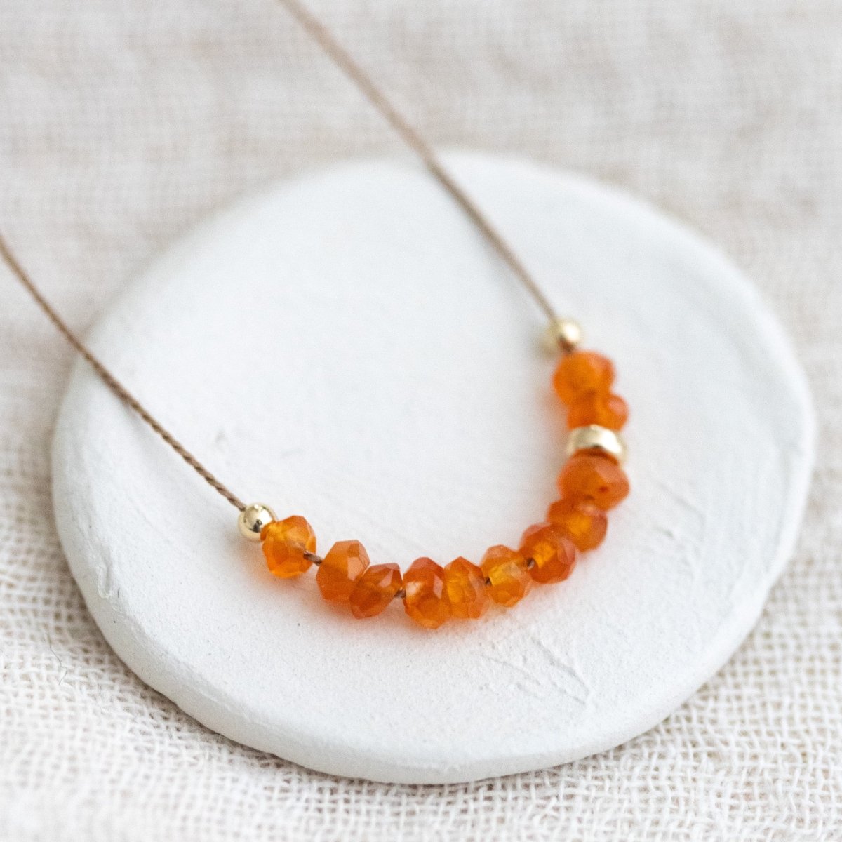 Auf einem weißen Steinschälchen liegt eine dünne, beige Schnurkette, auf die eine Reihe oranger Karneolperlen und einige goldene Akzentperlen auf gezogen sind. Weißer Stoff als Untergrund.