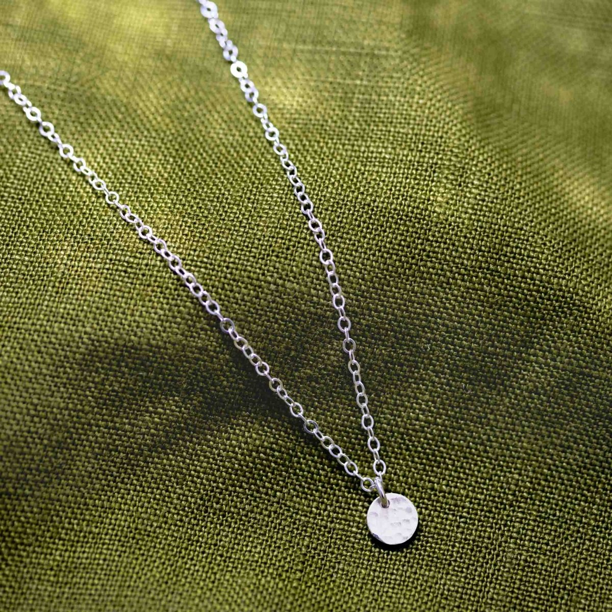 Eine feine Ankerkette aus 925 Silber mit einem kleinen, gehämmerten Plättchenanhänger liegt auf einem grünen Hintergrund.