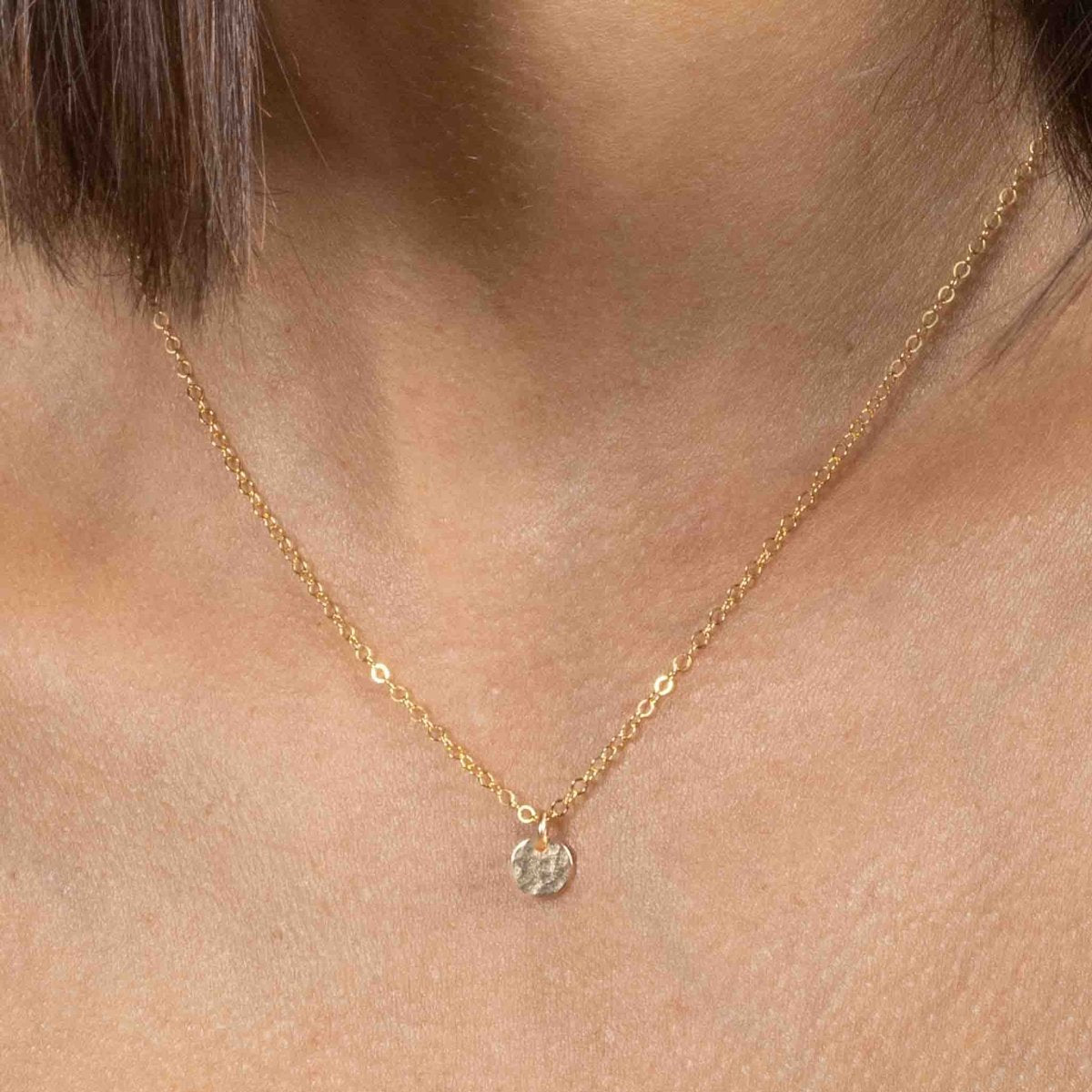 Am Hals des Models hängt eine kurze, goldene Ankerkette mit einem kleinen, gehämmerten Plättchen-Anhänger.