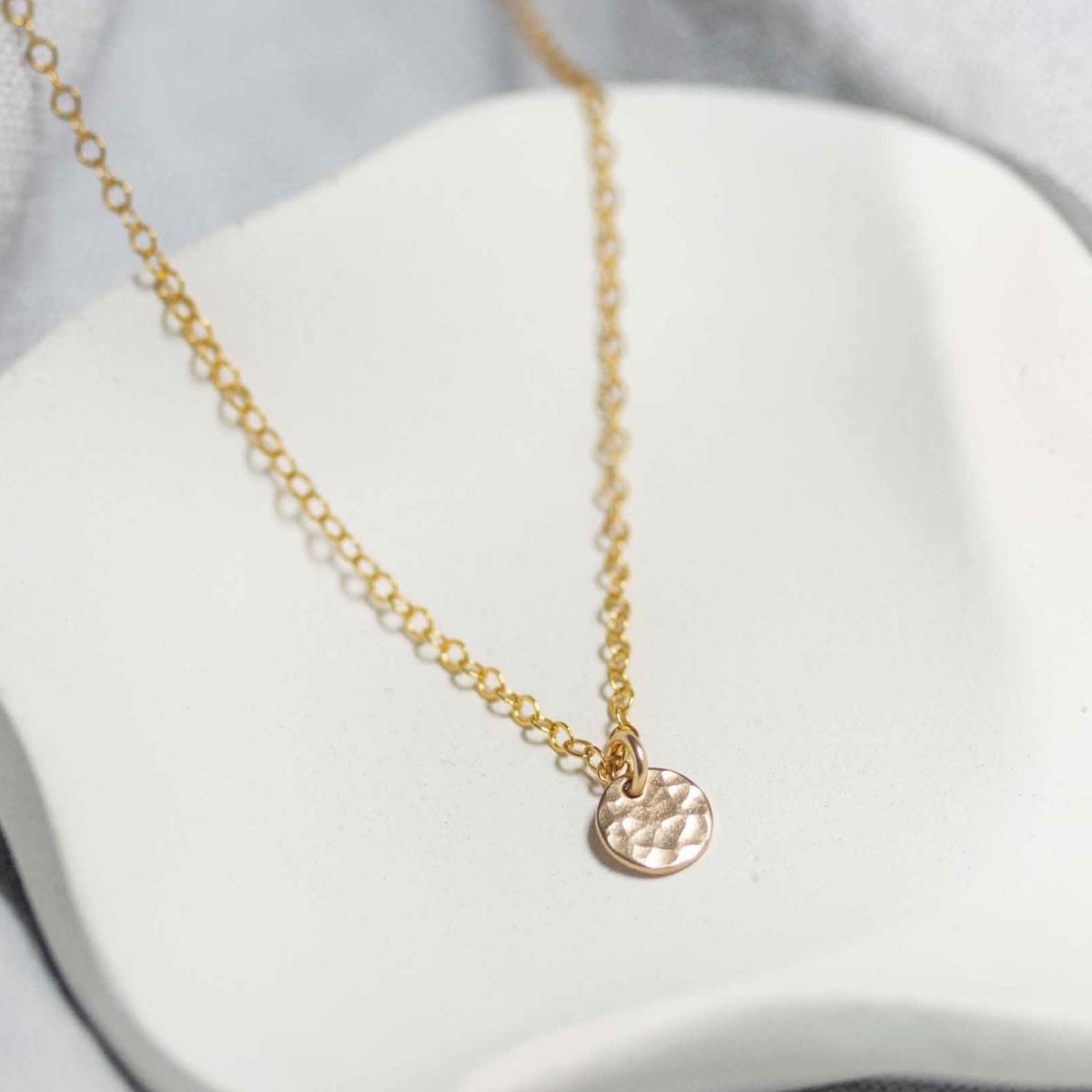 Eine goldene Ankerkette mit einem kleinen, gehämmerten Plättchen-Anhänger liegt auf einem weißen Schälchen.