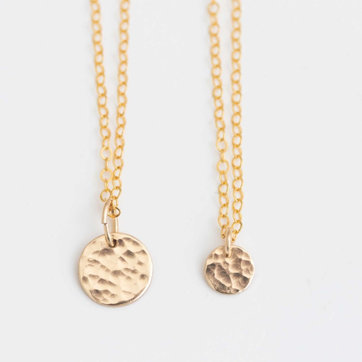 Zwei goldene Halsketten hängen vor weißem Hintergrund. An der einen Kette hängt ein 9 mm Plättchen, an der anderen ein 6 mm Plättchen, beide gehämmert.