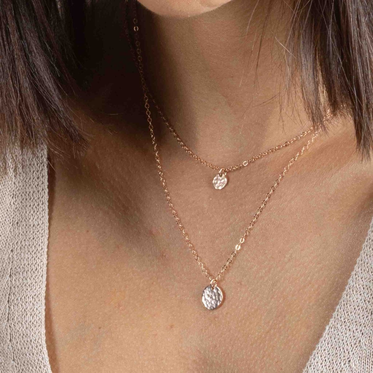 Das Model trägt zwei rosé goldene Halsketten: an der kürzeren hängt ein kleines, gehämmertes Plättchen, an der längeren ein etwas größeres, ebenfalls gehämmertes Plättchen.