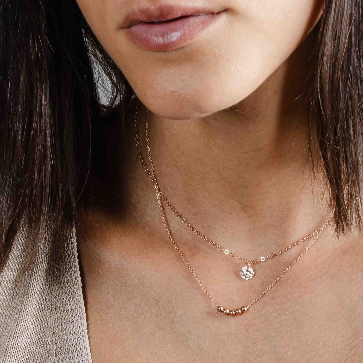 Das Model trägt zwei rosegoldene Ketten im Layer Look: an der kürzeren Kette hängt ein kleines, gehämmertes Plättchen, auf die längere Kette sind 5 rosé goldene Perlen aufgezogen.