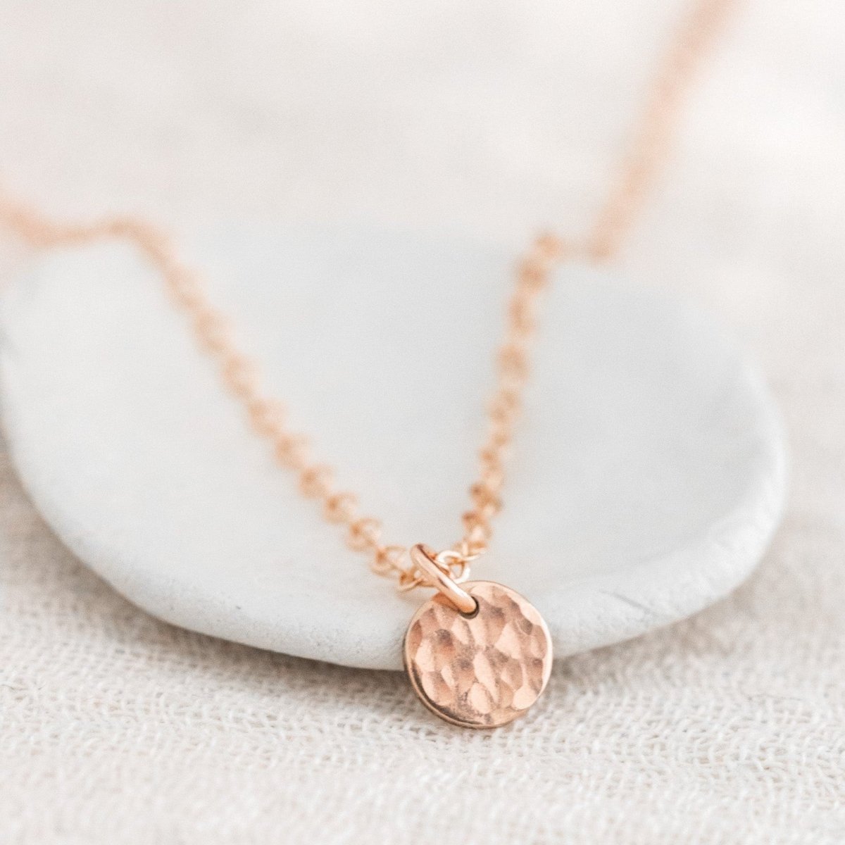Eine feine rosé goldene Halskette mit einem kleinen, gehämmerten Plättchen lehnt an einem weißen Schälchen auf weißem Untergrund.