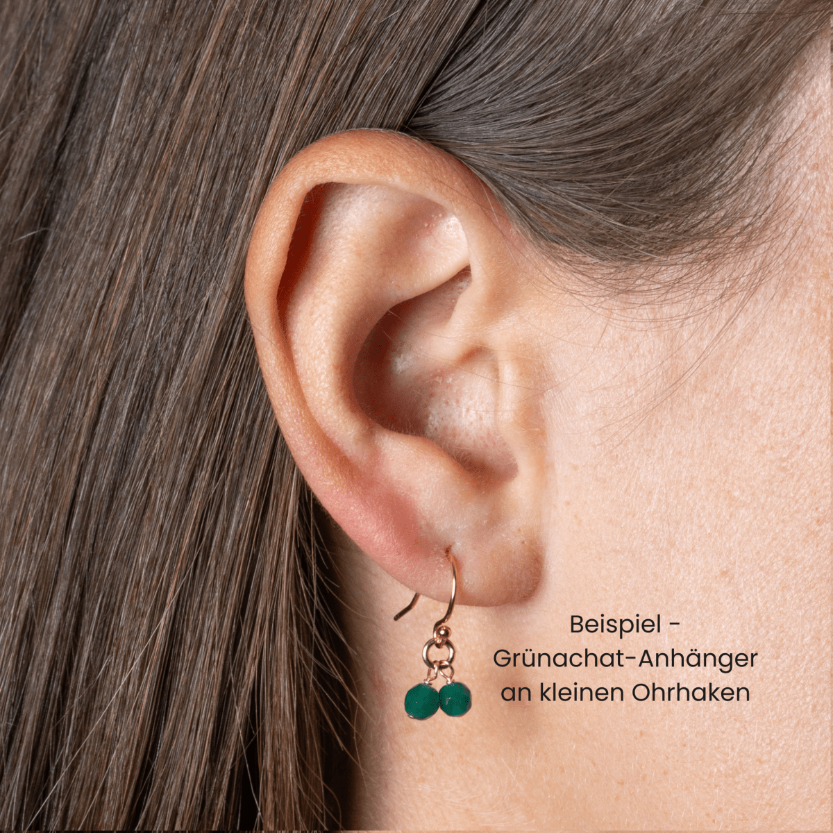 Am Ohr des Models hängt ein Geburtsstein-Ohrring, der aus einem goldenen Ohrhaken und zwei kleinen Achat-Edelsteinen besteht. Die deutsche Beschriftung daneben lautet: Beispiel - Grünachat-Anhänger an kleinen Ohrhaken.