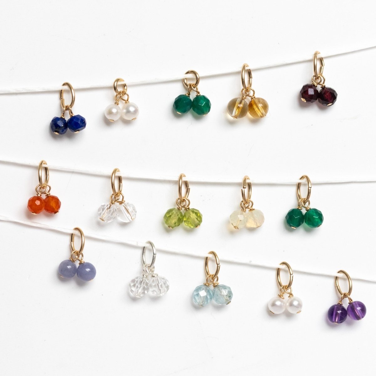 Vor einem weißen Hintergrund hängen drei Reihen kleiner Geburtsstein-Anhänger. Jeder Anhänger besteht aus zwei runden Edelsteinen an einem kleinen Ring.