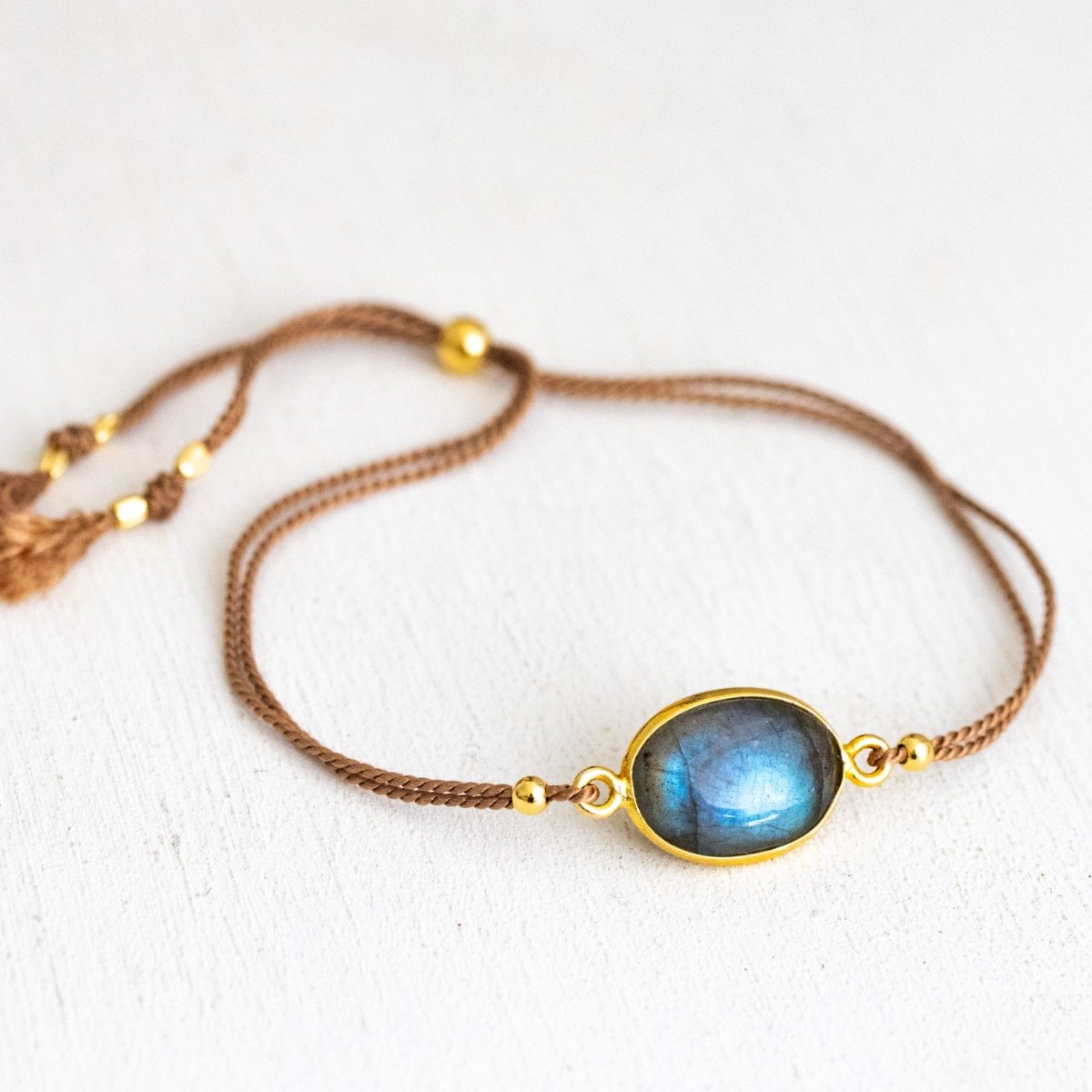Ein dünnes, beiges Seidenarmband mit einem ovalen und in gold gefassten Labradorit in der Mitte liegt auf weißem Hintergrund.