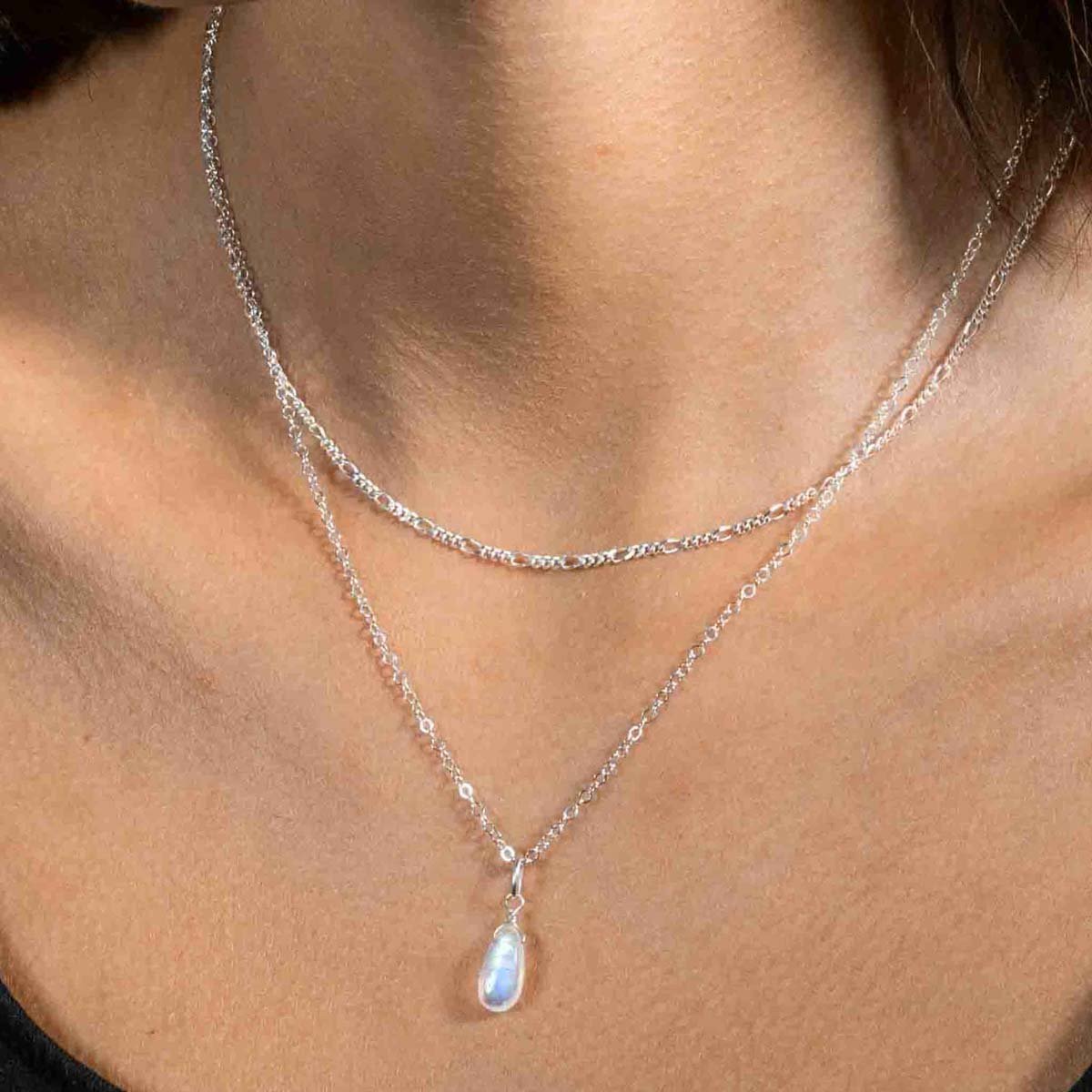 Am Hals des Models hängt eine kürzere Figaro-Kette aus Silber und eine längere Ankerkette aus Silber mit einem tropfenförmigen Mondstein-Anhänger.