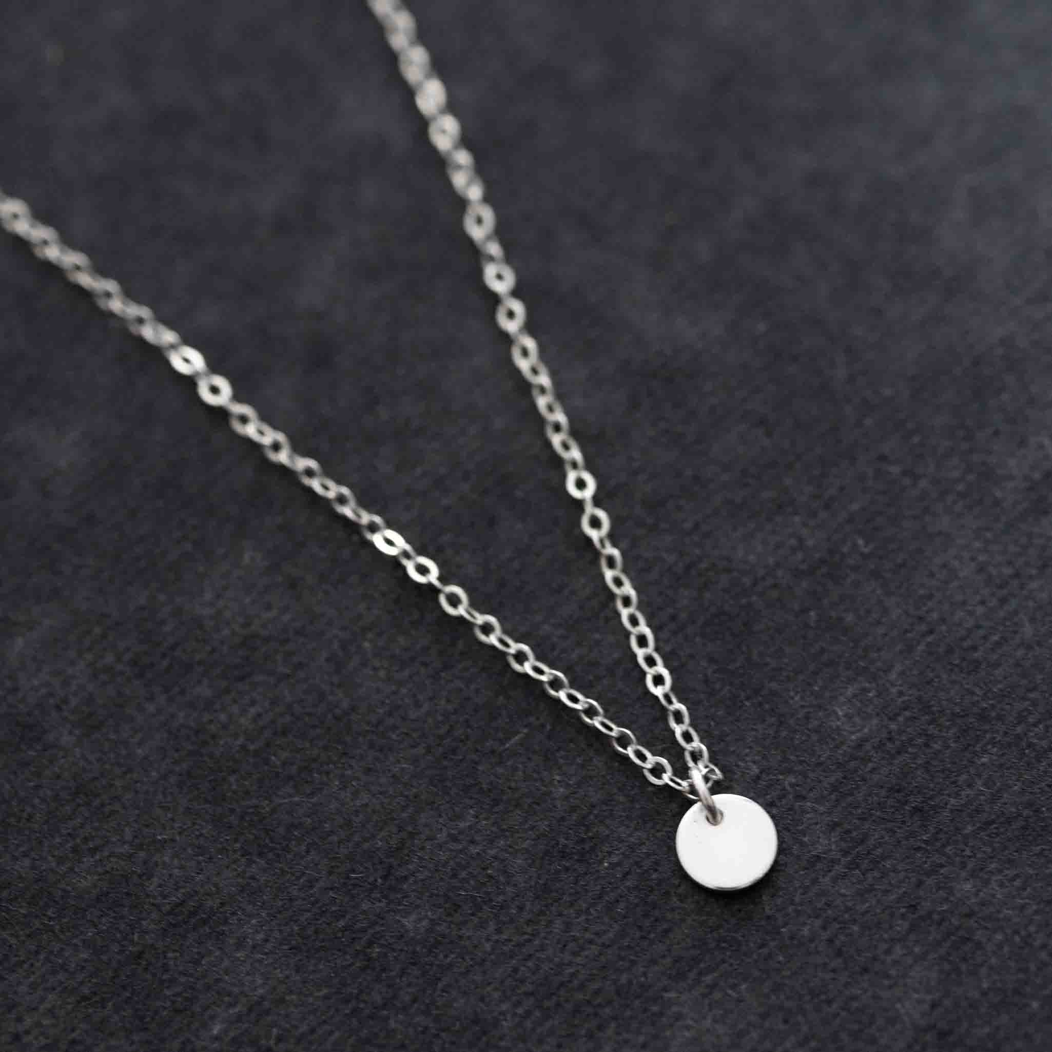 Eine zierliche Ankerkette aus 925 Silber mit einem glänzenden, runden Plättchen-Anhänger liegt auf grauem Hintergrund.