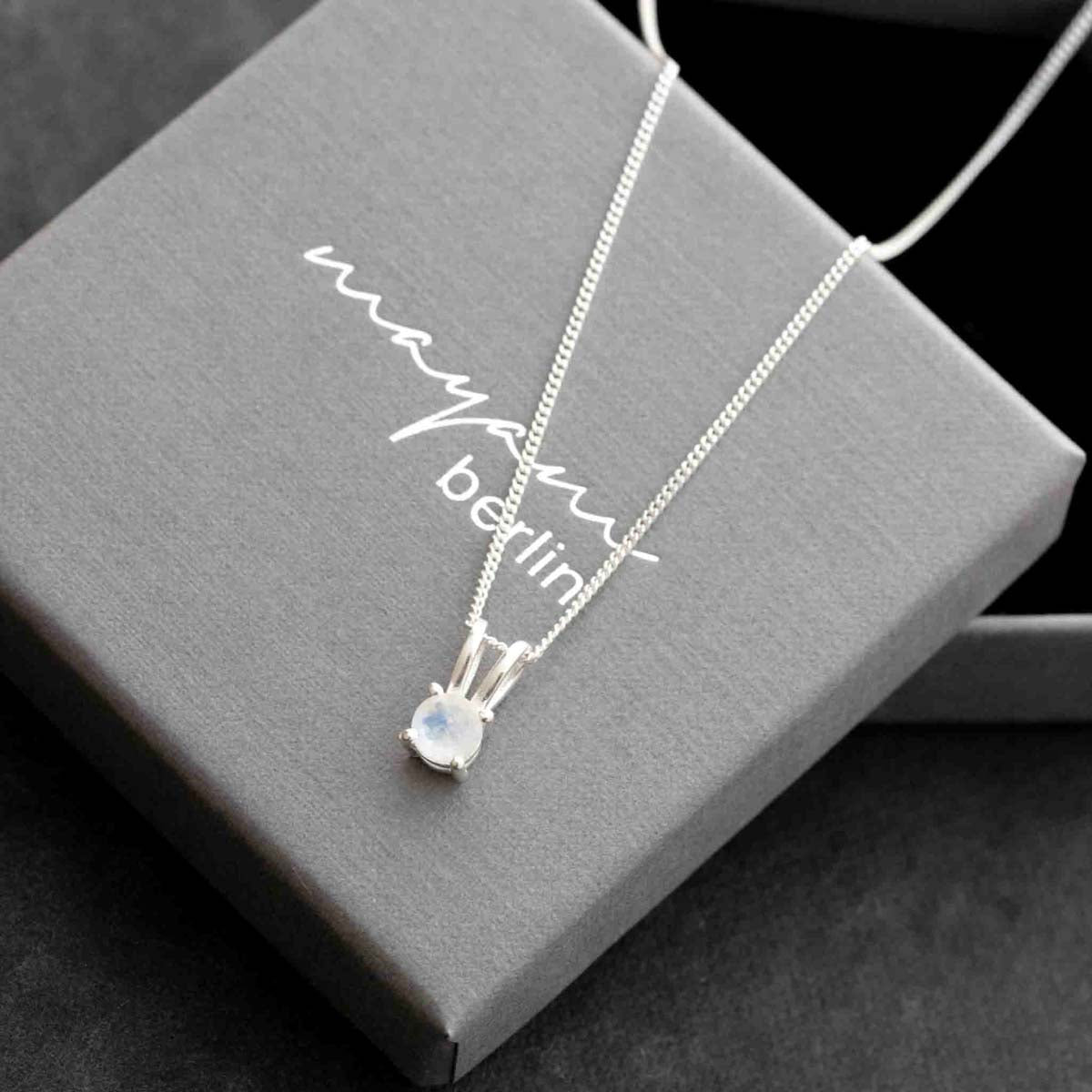 Eine silberne Halskette mit einem kleinen, runden Mondstein-Anhänger liegt auf einem grauen Schmucketui.