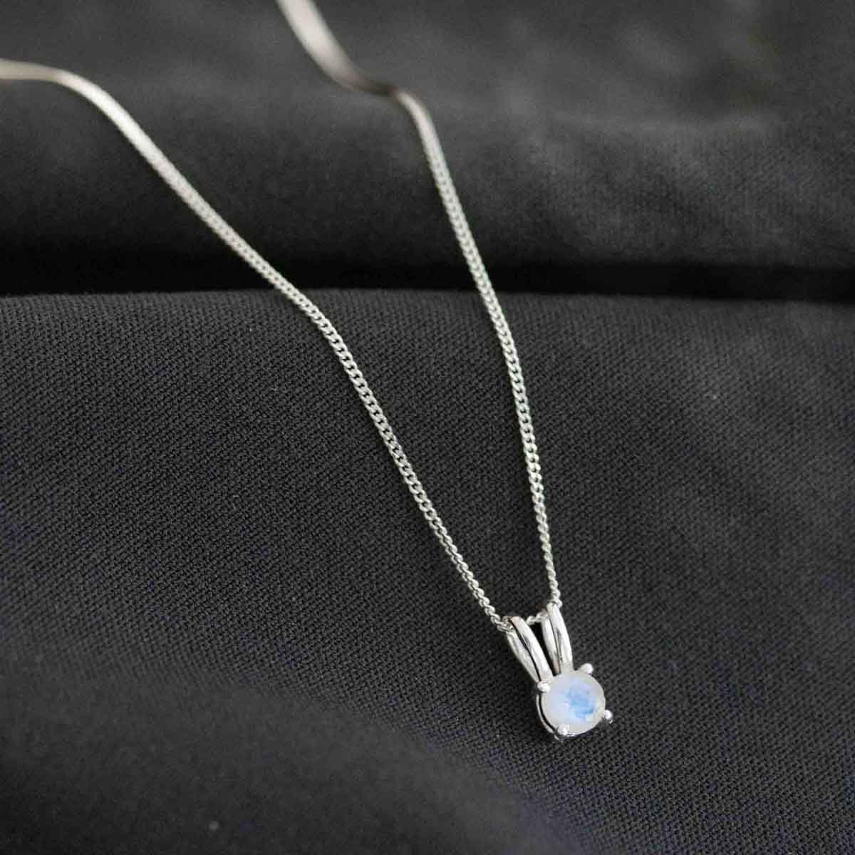 Eine feine, silberne Panzerkette mit einem winzigen, runden, in Silber gefassten Mondstein Anhänger auf dunklem Hintergrund.