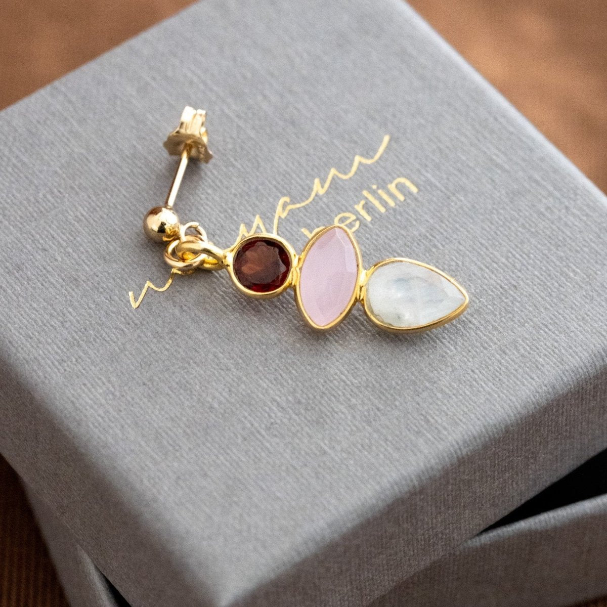 Ein goldener Multi-Edelstein-Ohrstecker, bestehend aus Granat, rosa Chalcedon und Mondstein, liegt auf einem grauen Schmucketui.
