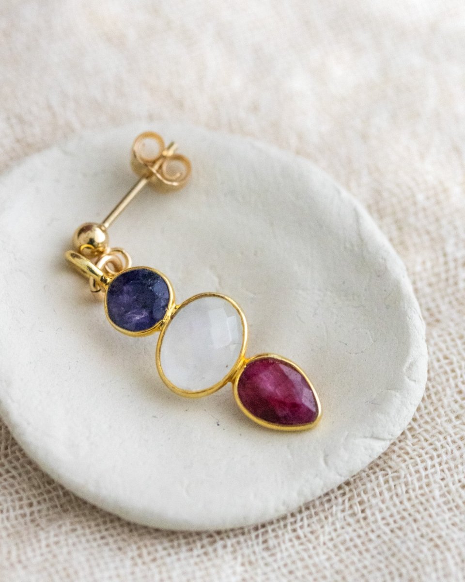 Ein Multi-Edelstein Ohrring mit Saphir, Mondstein und Rubin in gold gefasst liegt in einem weißen Schälchen auf naturfarbenen Leinenstoff.