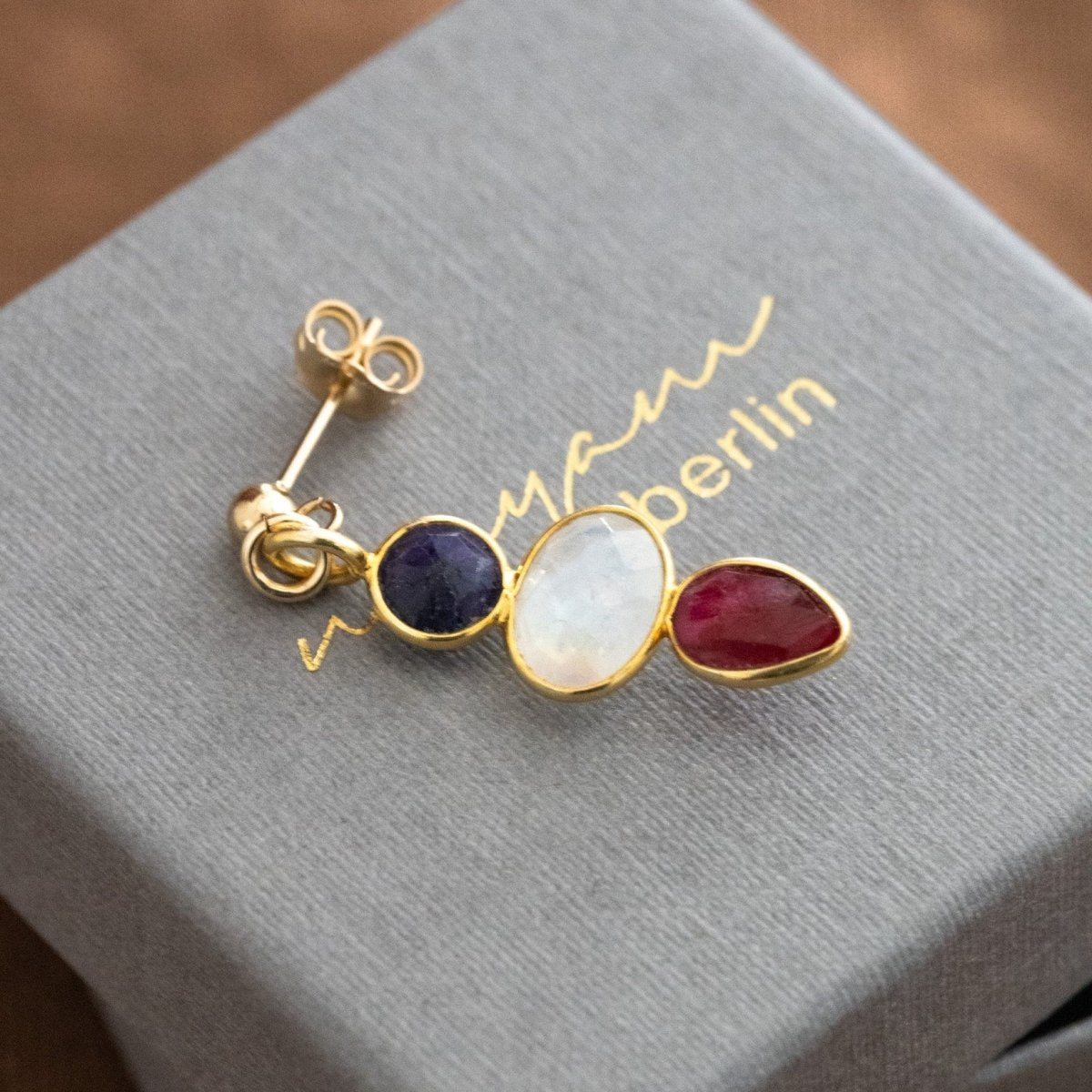 Ein Multi-Edelstein Ohrring mit Saphir, Mondstein und Rubin in gold gefasst liegt auf einem grauen Schmucketui.
