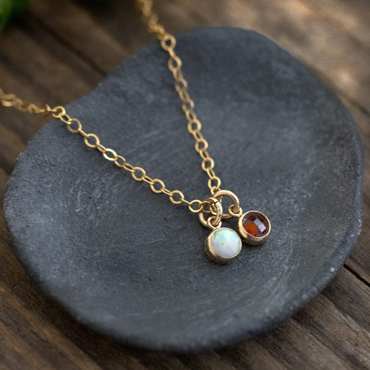Eine zierliche Goldkette mit zwei winzigen, runden Anhängern, einem Opal und einem Granat, auf einem grauen Steinschälchen, das auf Holzuntergrund steht.