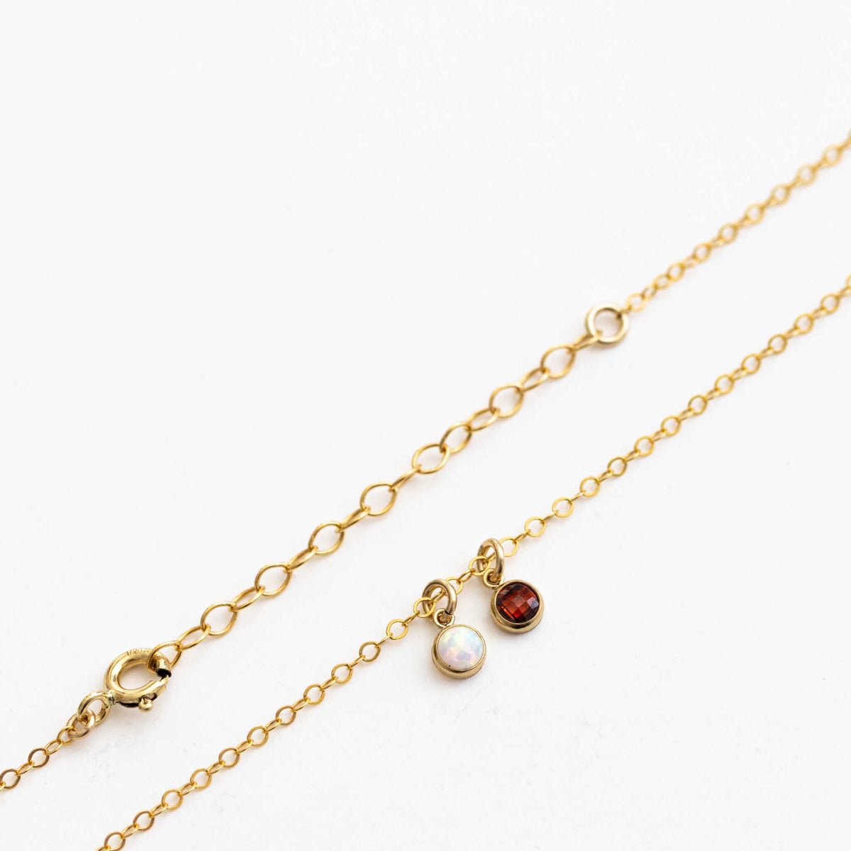 Eine zierliche Goldkette mit zwei winzigen, runden Anhängern, einem Opal und einem Granat, auf weißem Hintergrund.