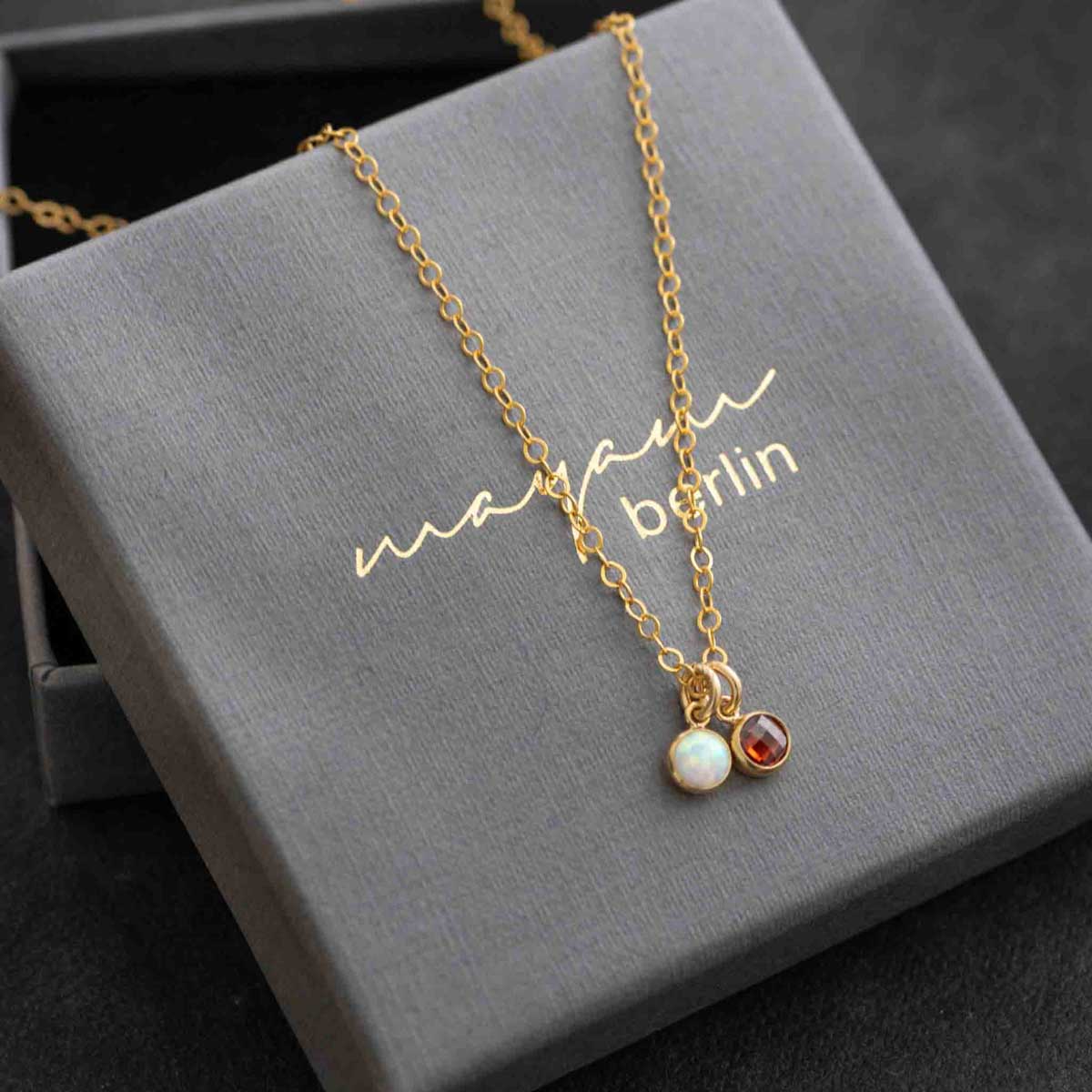Eine zierliche Goldkette mit zwei winzigen, runden Anhängern, einem Opal und einem Granat, auf einem grauen Schmucketui, beschriftet mit "mayam berlin".