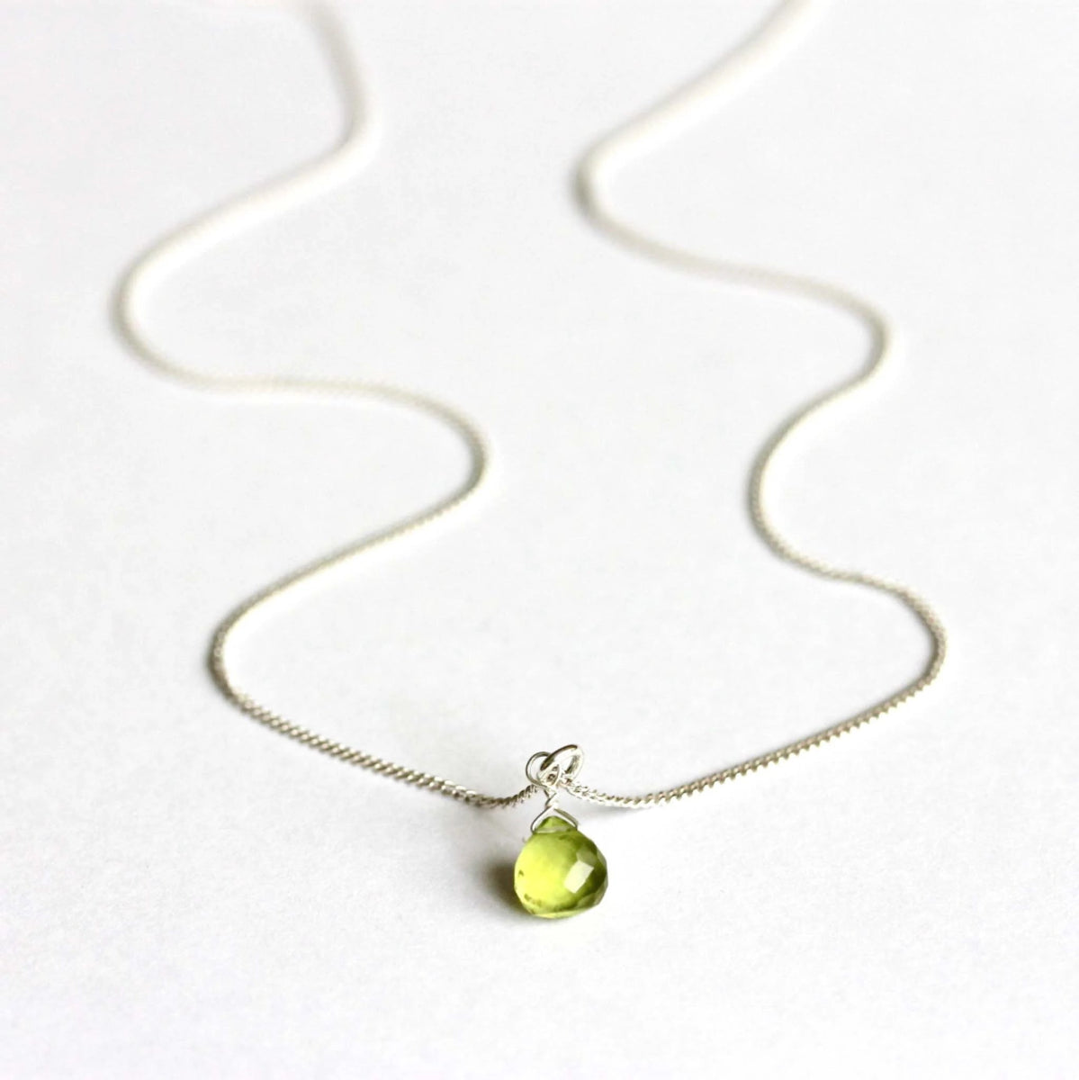 Eine feine 925 Silberkette mit einem kleinen, tropfenförmigen Peridot-Anhänger liegt auf weißem Hintergrund.