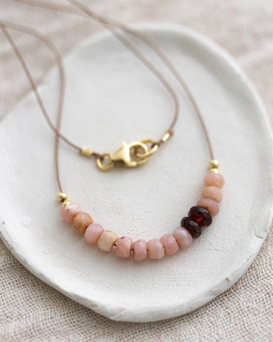 Eine Halskette aus dünner, beiger Seidenschnur, auf die eine Reihe rosa Opale und dunkelroter Granate aufgezogen sind, liegt auf einem weißen Schälchen.