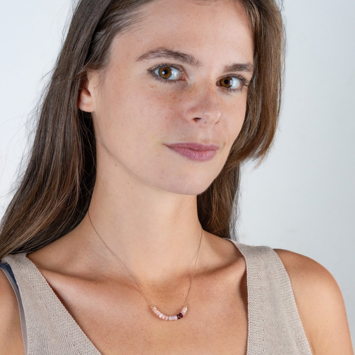 Am Hals des Models hängt eine kurze Halskette, die aus einer dünnen, beigen Seidenschnur besteht, auf die eine kurze Reihe rosa Opale und zwei schwarzrote Granate aufgezogen sind.