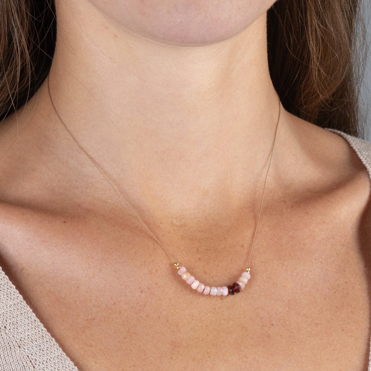 Am Hals des Models hängt eine kurze Halskette, die aus einer dünnen, beigen Seidenschnur besteht, auf die eine kurze Reihe rosa Opale und zwei schwarzrote Granate aufgezogen sind.