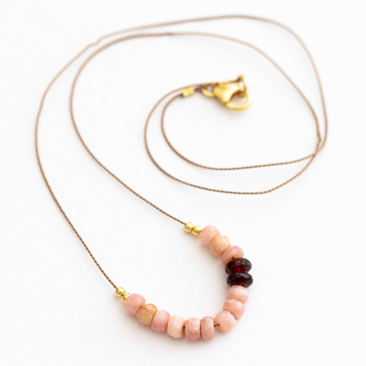 Eine Halskette aus dünner, beiger Seidenschnur, auf die eine Reihe rosa Opale und dunkelroter Granate aufgezogen sind, liegt auf weißen Hintergrund.
