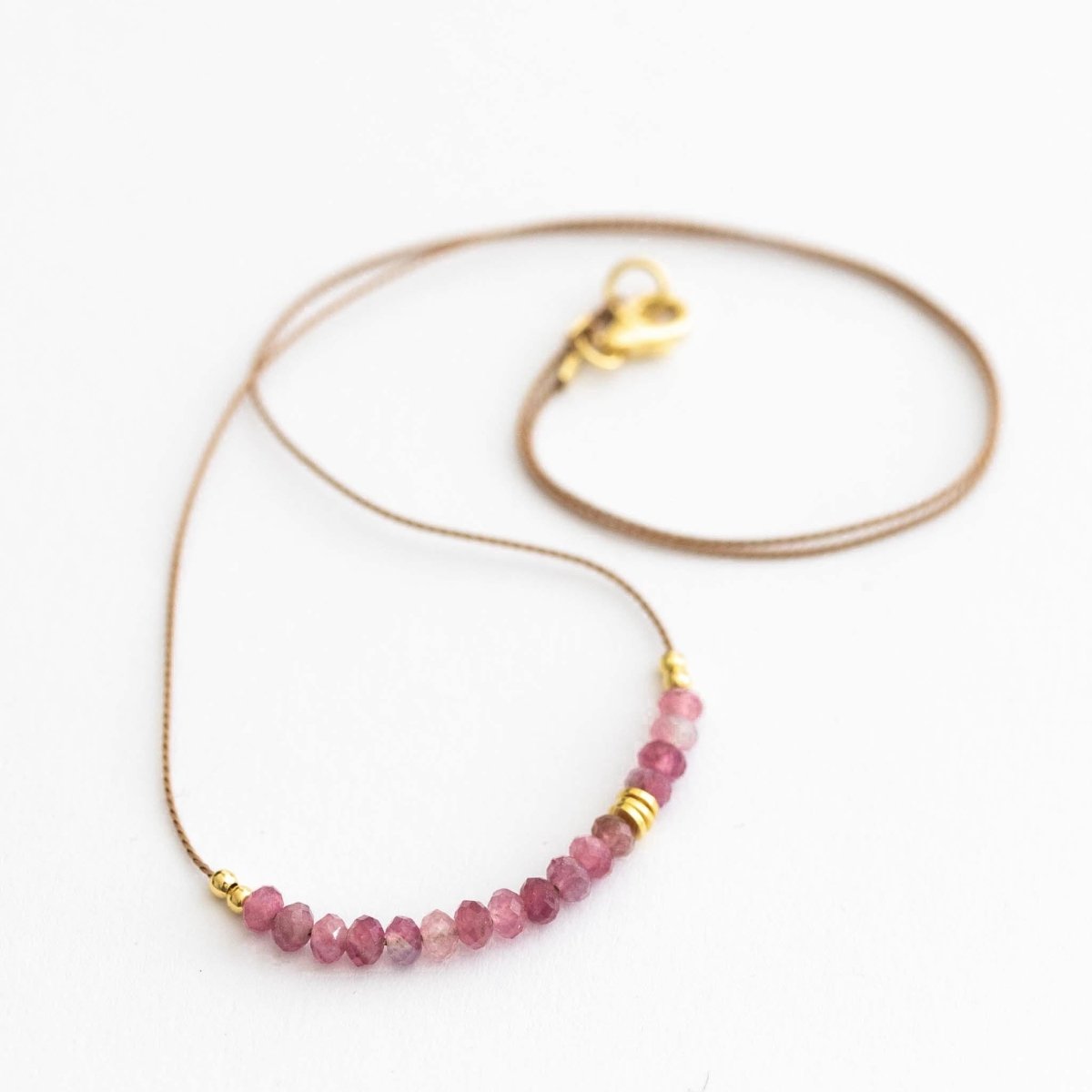 Auf weißen Hintergrund liegt eine kurze Halskette. Sie besteht aus einer dünnen Seidenschnur mit einer Reihe kleiner, rosa Turmalin-Rondelle und goldenen Akzenten in der Mitte.