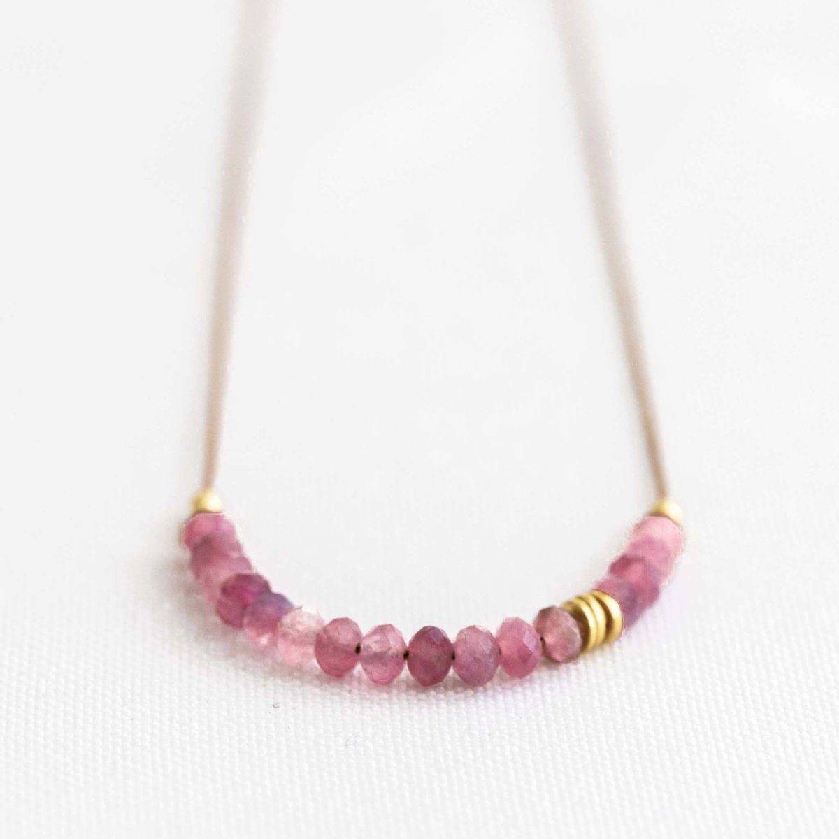 Auf weißen Hintergrund liegt eine kurze Halskette. Sie besteht aus einer dünnen Seidenschnur mit einer Reihe kleiner, rosa Turmalin-Rondelle und goldenen Akzenten in der Mitte.