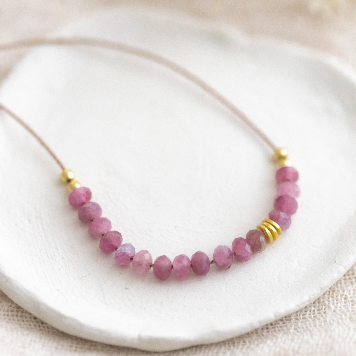 Auf weißen Schälchen liegt eine kurze Halskette. Sie besteht aus einer dünnen Seidenschnur mit einer Reihe kleiner, rosa Turmalin-Rondelle und goldenen Akzenten in der Mitte.
