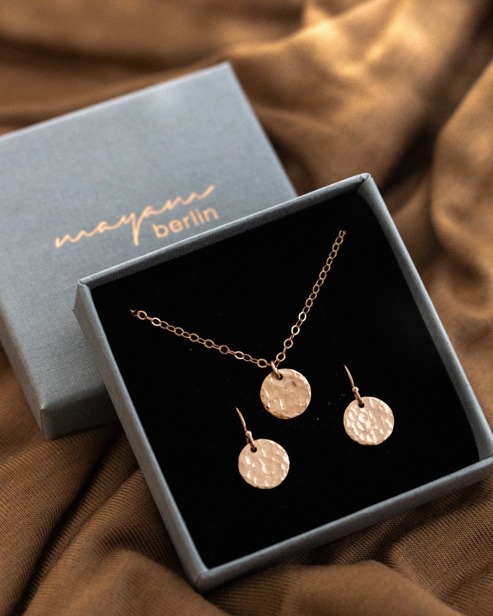 In einem geöffneten Schmucketui sind eine feine Rosegold-Kette mit gehämmertem Plättchen-Anhänger und passenden Ohrringen in den Schaumstoff eingehängt.