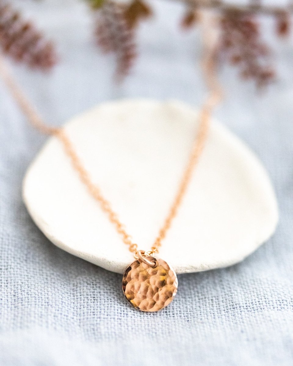 Eine feine Roségoldkette mit einem runden, gehämmerten Plättchen-Anhänger lehnt an einem weißen Schälchen auf grau-blauem Stoff.