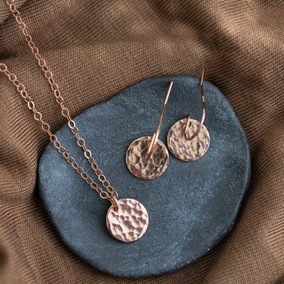 Auf einem grauen Steinschälchen liegt ein rose goldenes Schmuckset, das aus einer Kette und einem Paar Ohrringen besteht, an denen jeweils ein gehämmertes, rundes Plättchen hängt. Brauner Stoffuntergrund.
