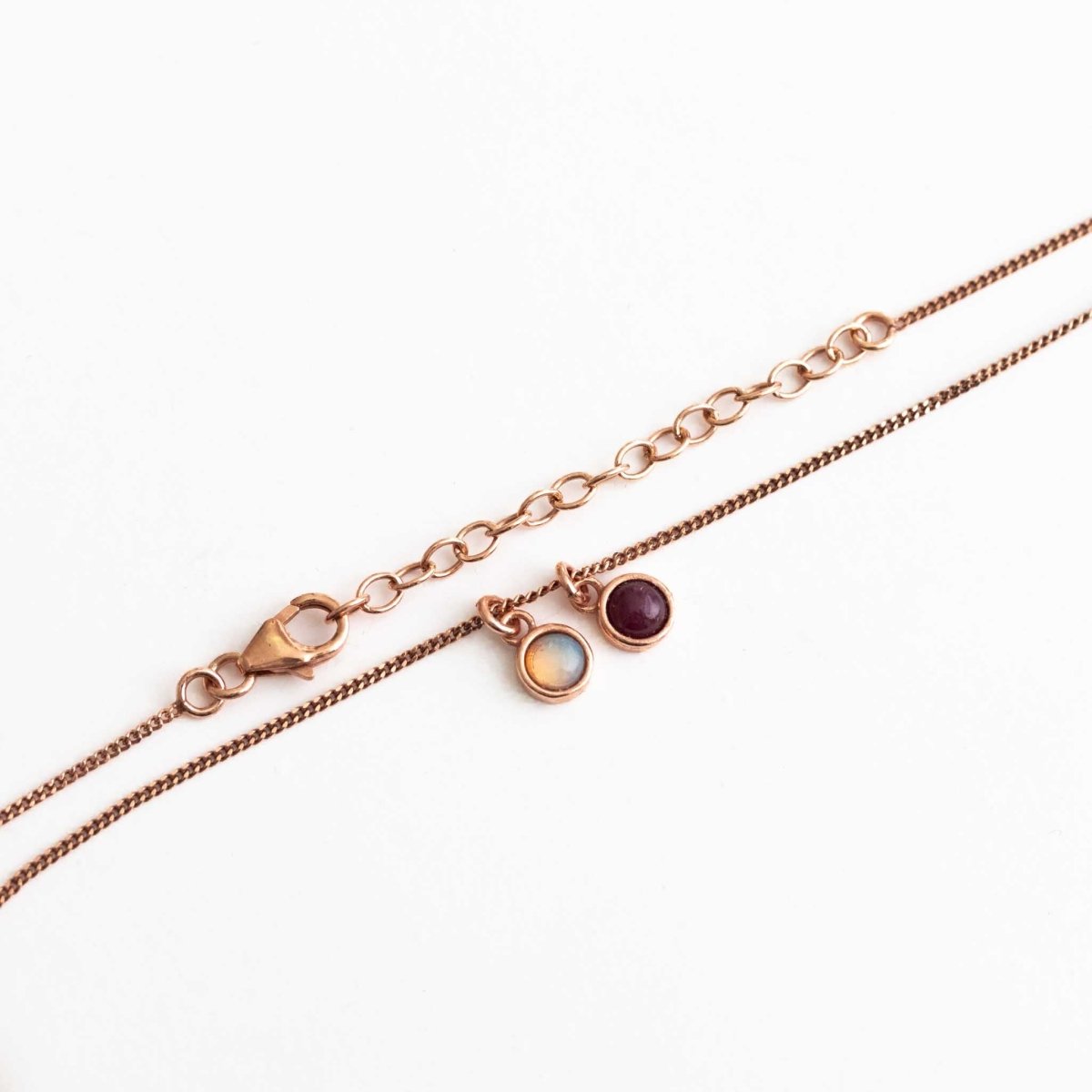 Eine feine Halskette aus rosé vergoldetem Silber mit zwei Edelstein-Anhängern, einem Opal und einem Rubin, auf weißem Hintergrund.