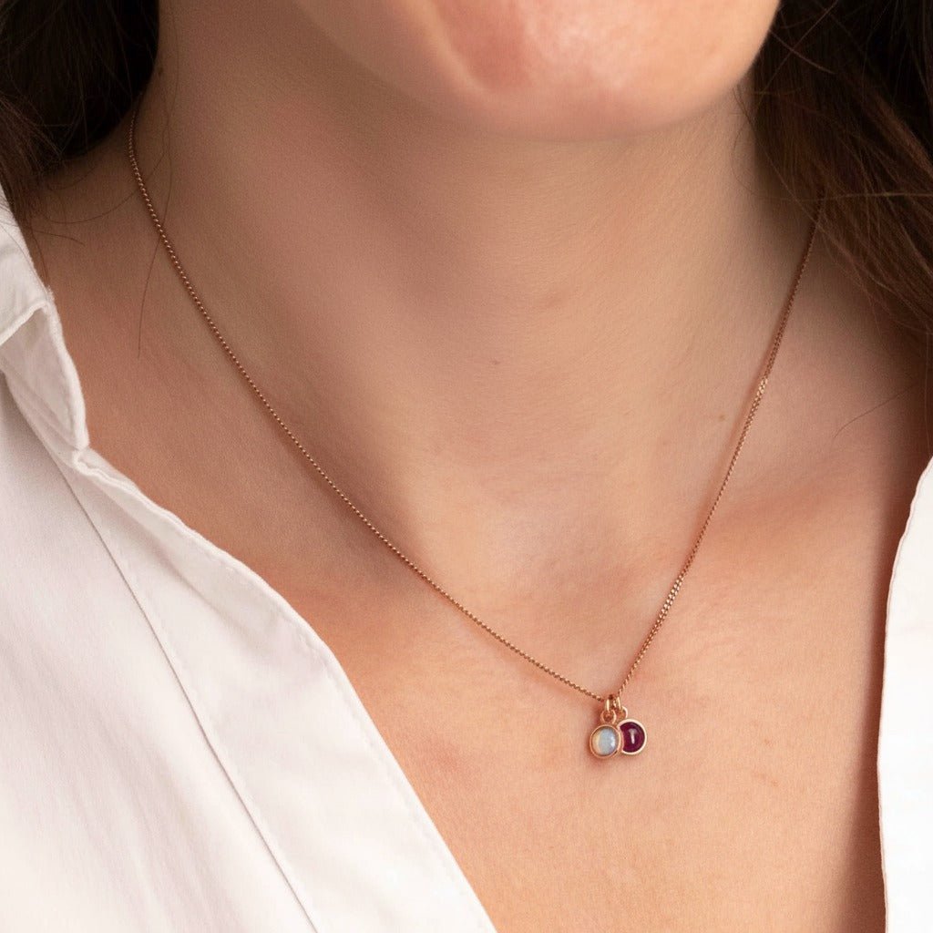 Eine rosé goldene Halskette mit einem winzigen Opal und einem winzigen Rubin-Anhänger am Hals des Models.
