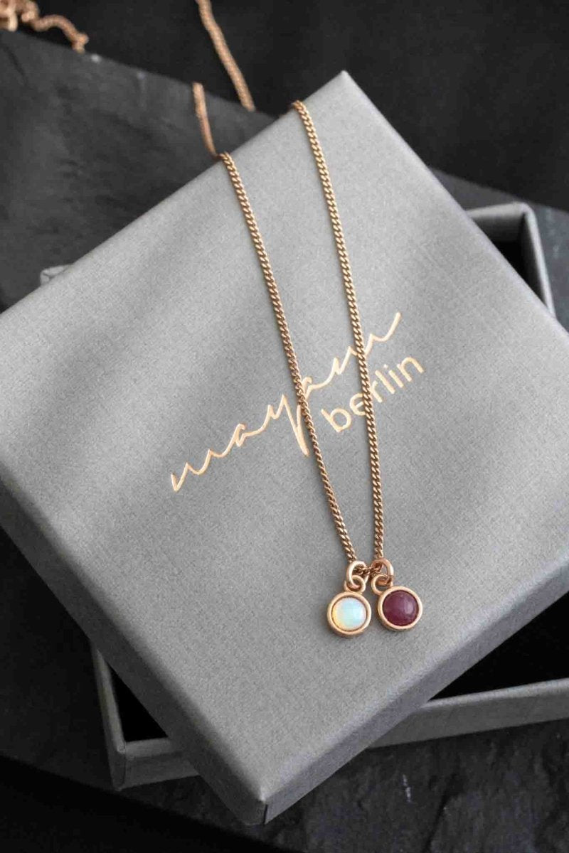 Eine feine Halskette aus rosé vergoldetem Silber mit zwei Edelstein-Anhängern, einem Opal und einem Rubin, auf einem grauen Schmucketui.