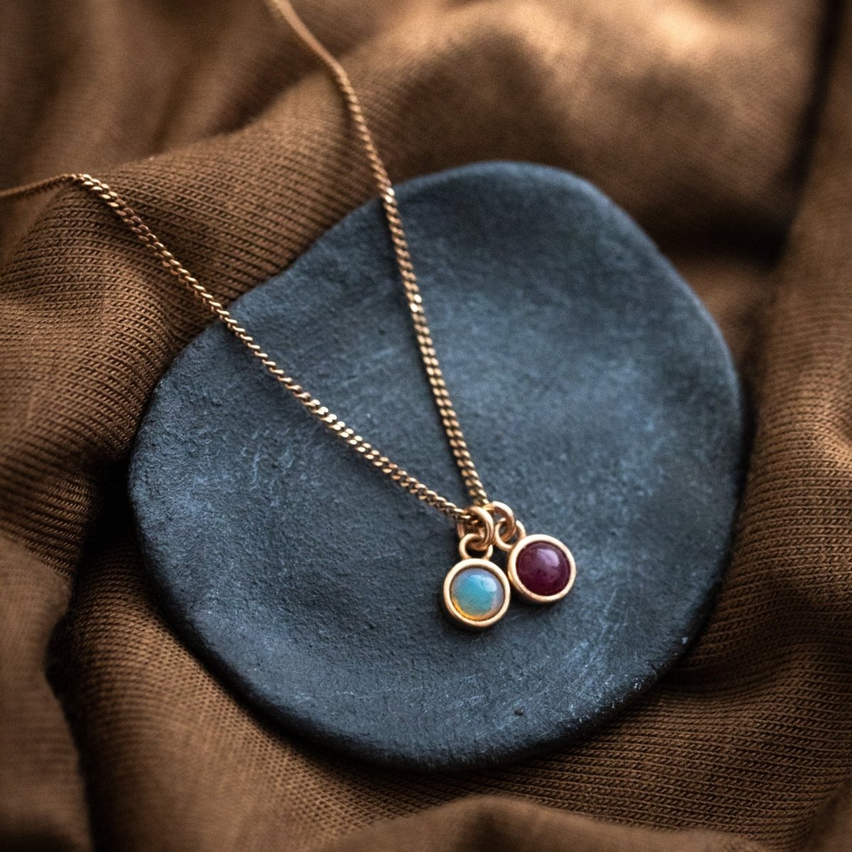 Eine feine Halskette aus rosé vergoldetem Silber mit zwei Edelstein-Anhängern, einem Opal und einem Rubin, auf einem grauen Schälchen auf braunem Stoff.