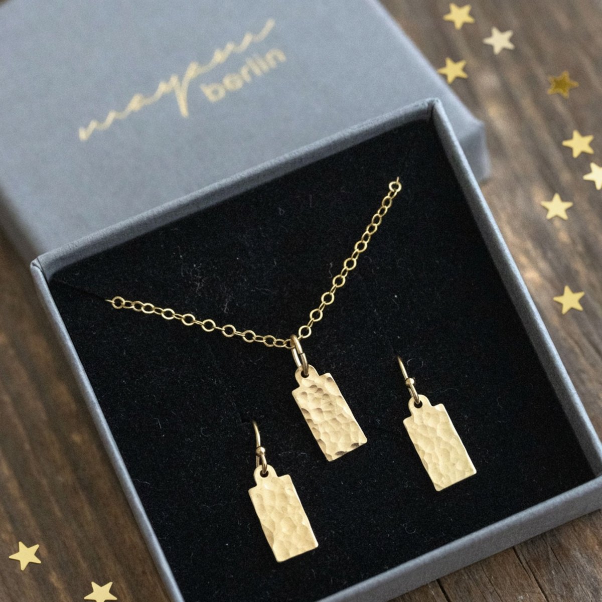 Im offenen Schmucketui ist ein Set eingehängt, bestehend aus einer feinen Goldkette und Ohrringen mit jeweils einem gehämmerten, rechteckigen Plättchen-Anhänger. Auf dem Holzuntergrund ist ein wenig goldenes Sternchen-Konfetti verteilt.