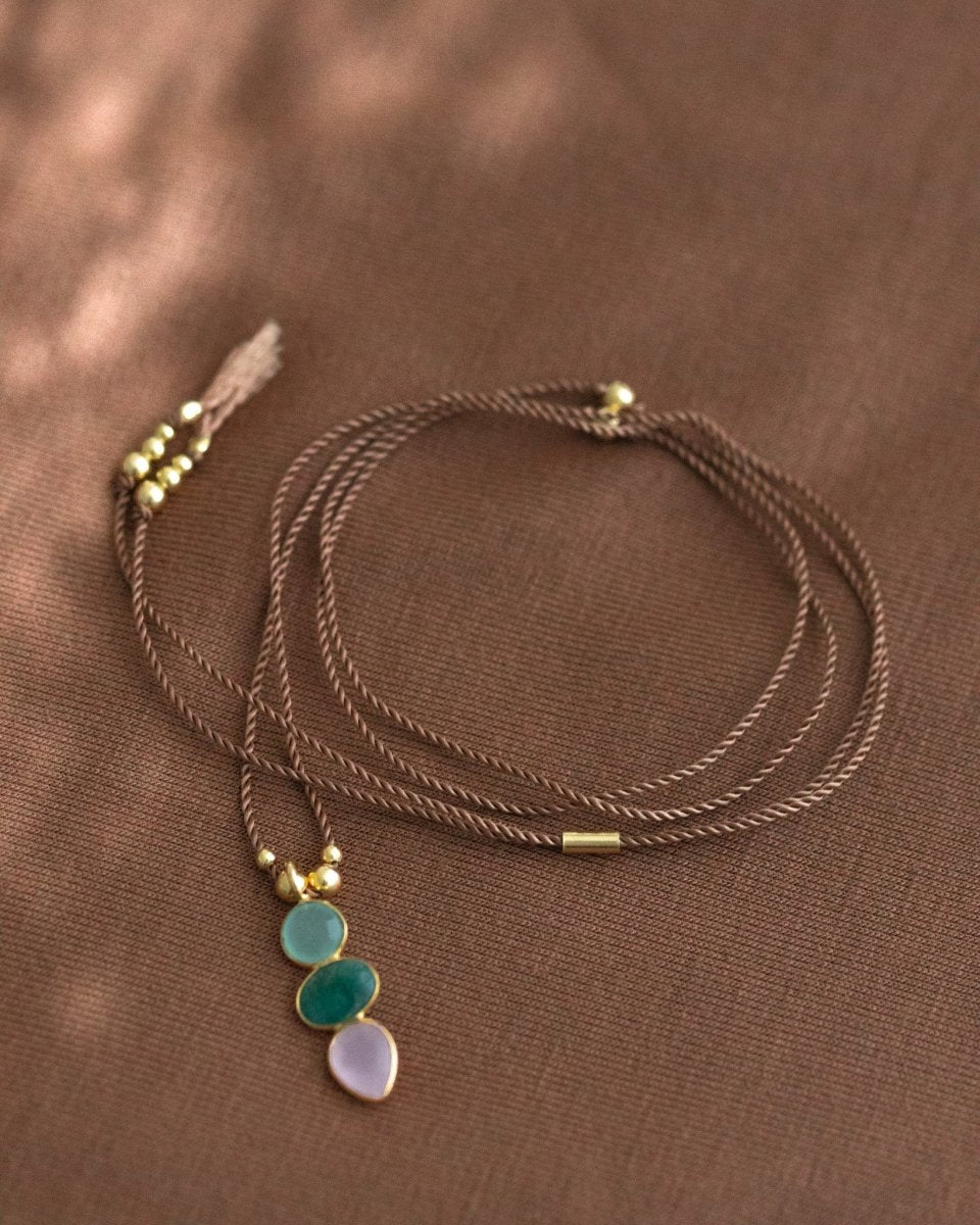 Eine dünne Schnurkette mit einem bunten Multi-Edelsteinanhänger und goldenen Zierelementen liegt auf braunem Hintergrund.