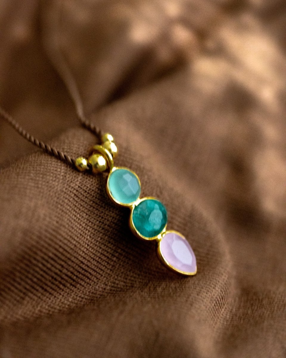 Eine dünne Schnurkette mit einem bunten Multi-Edelsteinanhänger und goldenen Zierelementen liegt auf braunem Hintergrund.