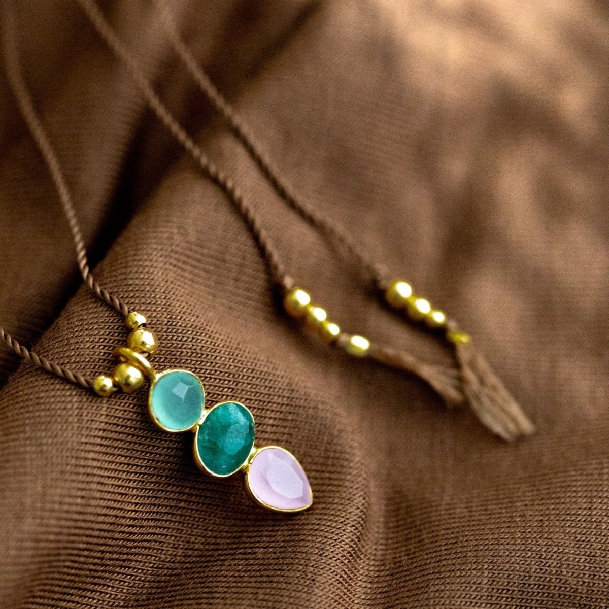 Eine dünne Schnurkette mit einem bunten Multi-Edelsteinanhänger und goldenen Zierelementen liegt auf braunem Hintergrund.