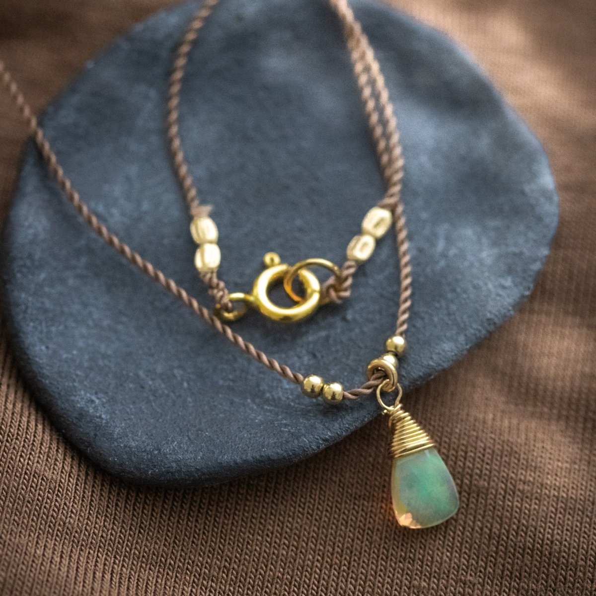 Eine dünne, braune Schnurkette mit einem dreieckigen Opal-Anhänger und goldenen Akzenten lehnt an einem schwarzen Schälchen.
