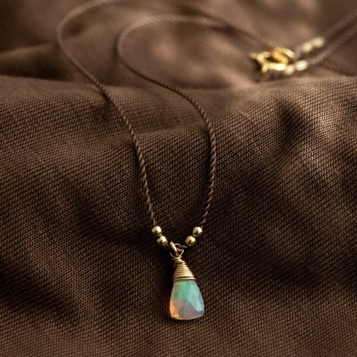 Eine dünne, braune Schnurkette mit einem dreieckigen Opal-Anhänger und goldenen Akzenten lehnt an braunem Stoff.