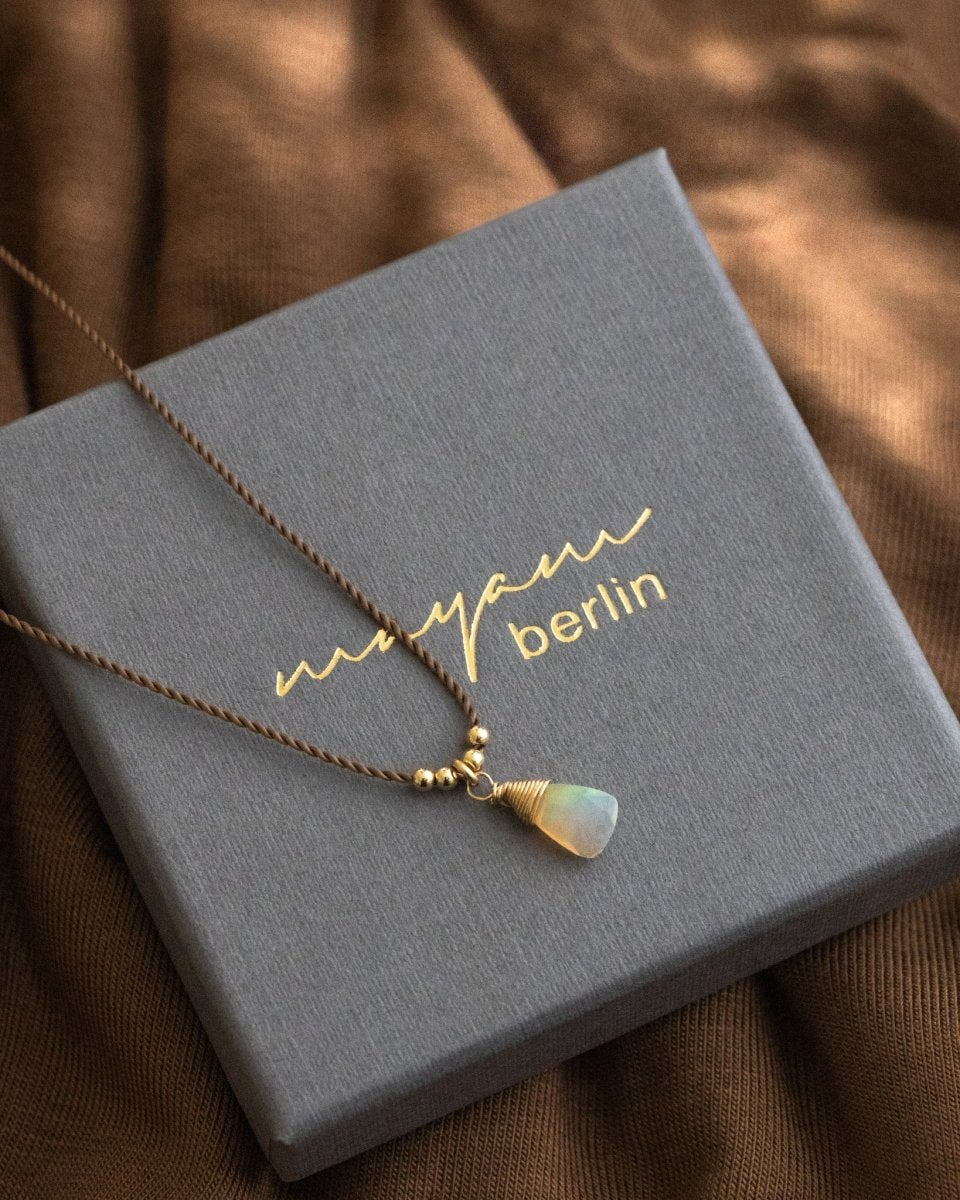 Eine dünne, braune Schnurkette mit einem dreieckigen Opal-Anhänger und goldenen Akzenten liegt auf einem grauen Schmucketui.
