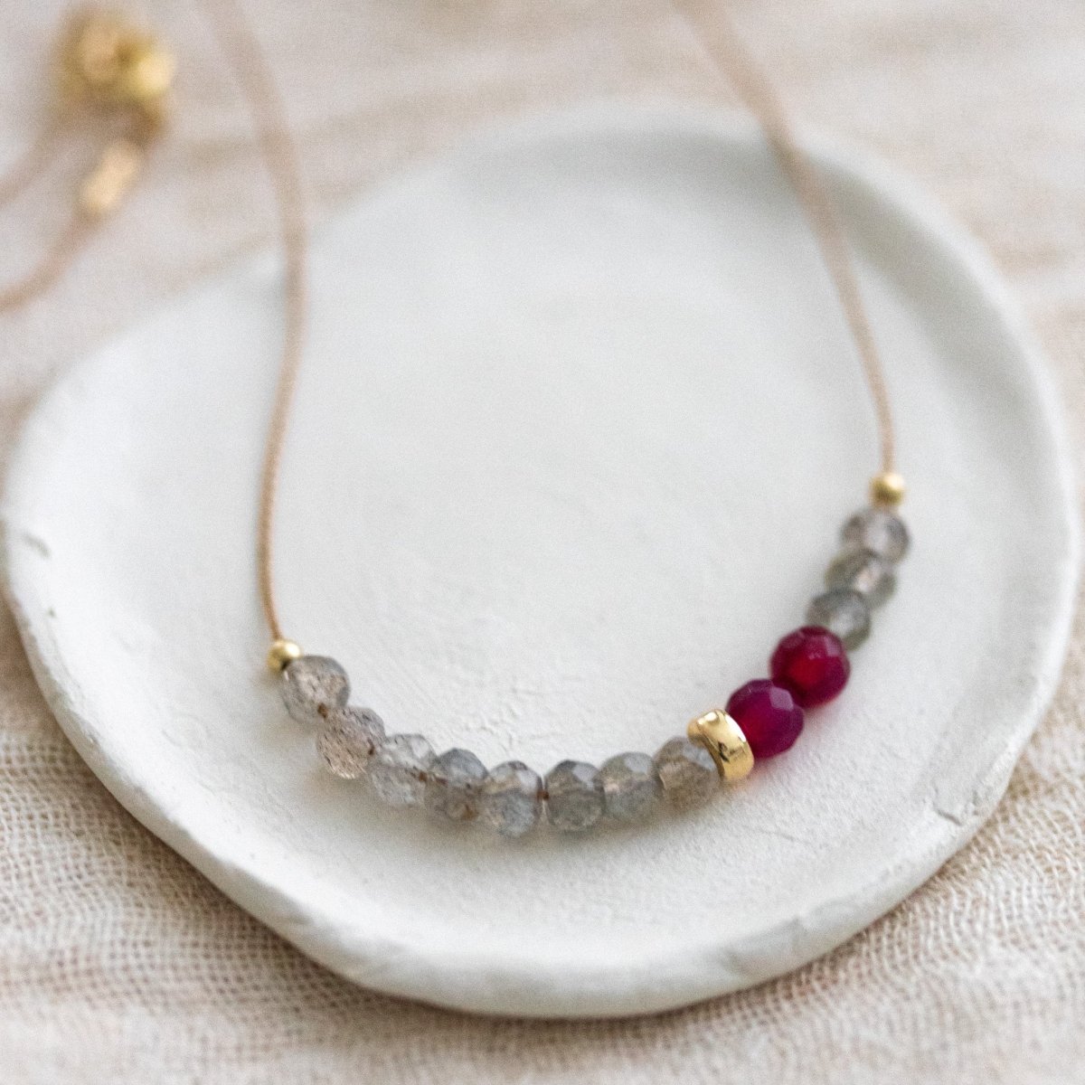 Eine Halskette aus beigem Seidenband, auf das einige Labradorit- und rote Achat-Rondelle aufgezogen sind, auf einem weißen Schälchen.